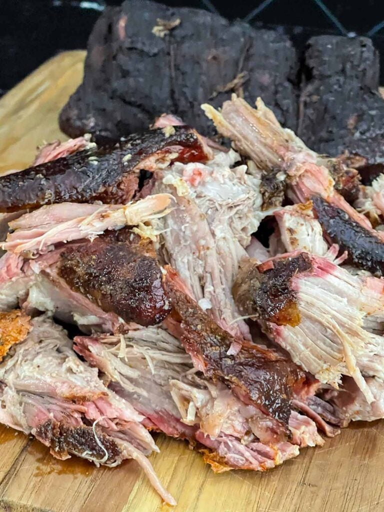 One smoked pork butt pulled apart beside the remaining whole half on a wooden cutting board.