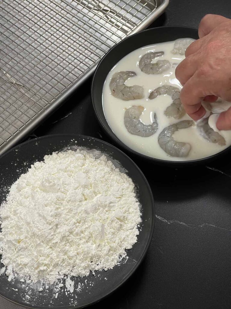 Raw shrimp soaking in buttermilk with a hand lifting one shrimp before dredging.