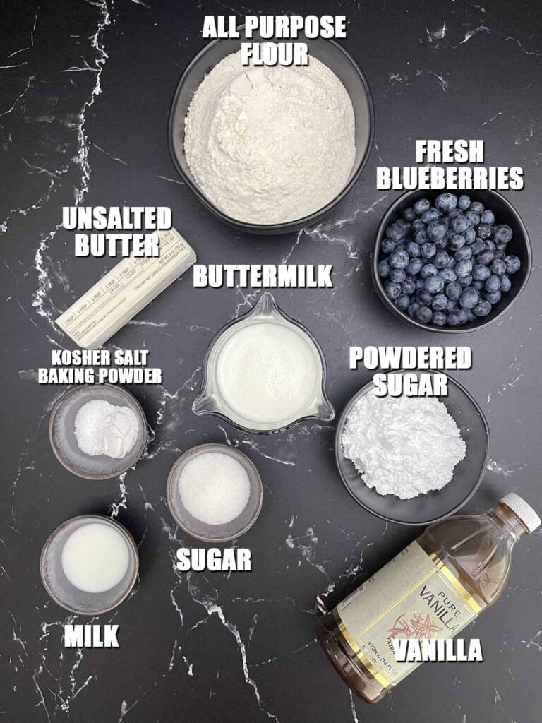 Ingredients for homemade blueberry biscuits including fresh blueberries, flour, butter, buttermilk, sugar, baking powder, salt, and vanilla glaze.