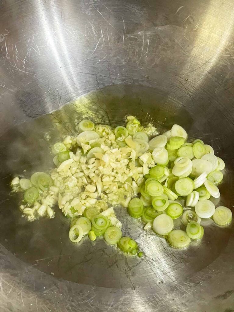 Finely chopped garlic and the white parts of green onions cooking in vegetable oil in a wok.