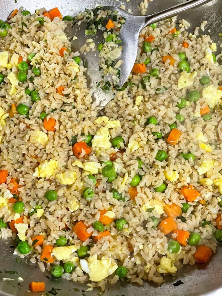 Brown rice, eggs, and vegetables tossed together in a wok with a large spatula.