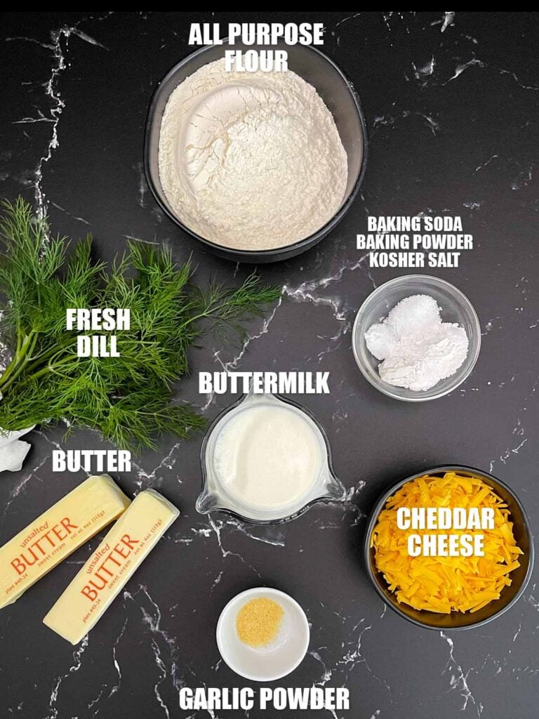 Ingredients for cheddar dill biscuits including flour, shredded cheddar cheese, fresh dill, buttermilk, butter, baking powder, baking soda, kosher salt, garlic powder, and butter for the topping.