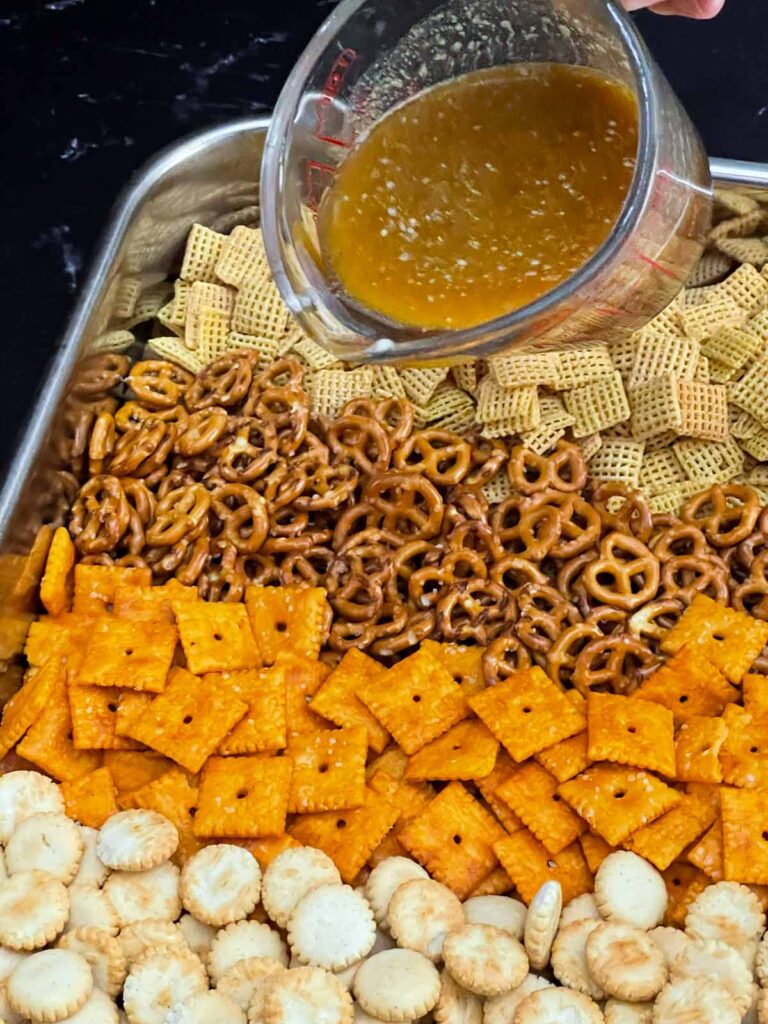 Old Bay butter mixture being poured over the snack mix ingredients in a roasting pan.
