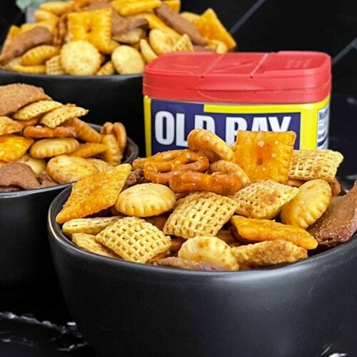 Old Bay Snack Mix served in a black bowl with Corn Chex cereal, pretzels, oyster crackers, cheddar crackers, and garlic rye chips.