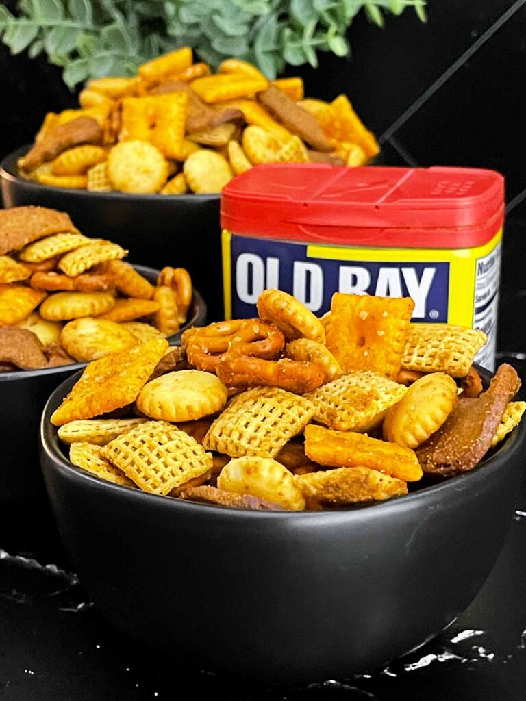 Old Bay Snack Mix served in a black bowl with Corn Chex cereal, pretzels, oyster crackers, cheddar crackers, and garlic rye chips.