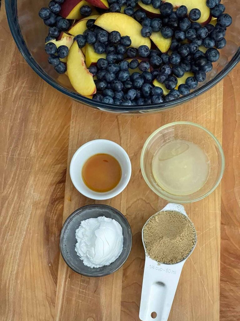 Peaches and blueberries in a mixing bowl with vanilla, lemon juice, cornstarch, and dark brown sugar in ramekins.