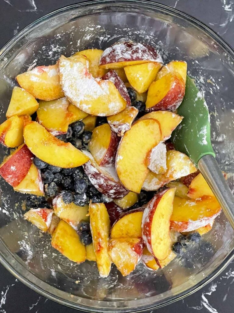 Fresh peaches and blueberries tossed with brown sugar, cornstarch, vanilla extract, and lemon juice in a large mixing bowl.
