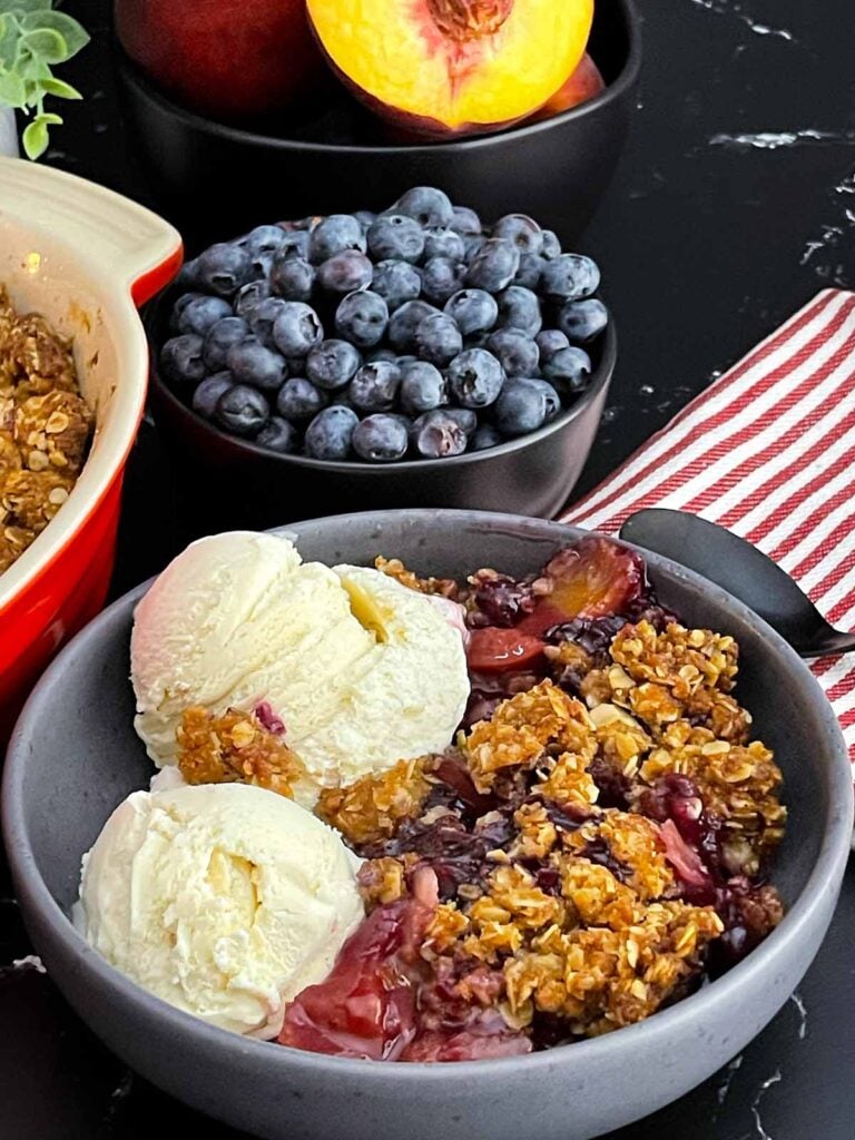 Golden brown Easy Peach Blueberry Crisp with a buttery oat topping served warm in a grey bowl with a scoop of vanilla ice cream melting over the fruit.
