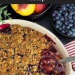 Easy Peach Blueberry Crisp with one serving scooped out, revealing the thick, jammy peach and blueberry filling beneath the golden oat topping, with a serving spoon resting in the baking dish.
