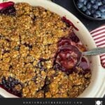 Easy Peach Blueberry Crisp with one serving scooped out, revealing the thick, jammy peach and blueberry filling beneath the golden oat topping, with a serving spoon resting in the baking dish.