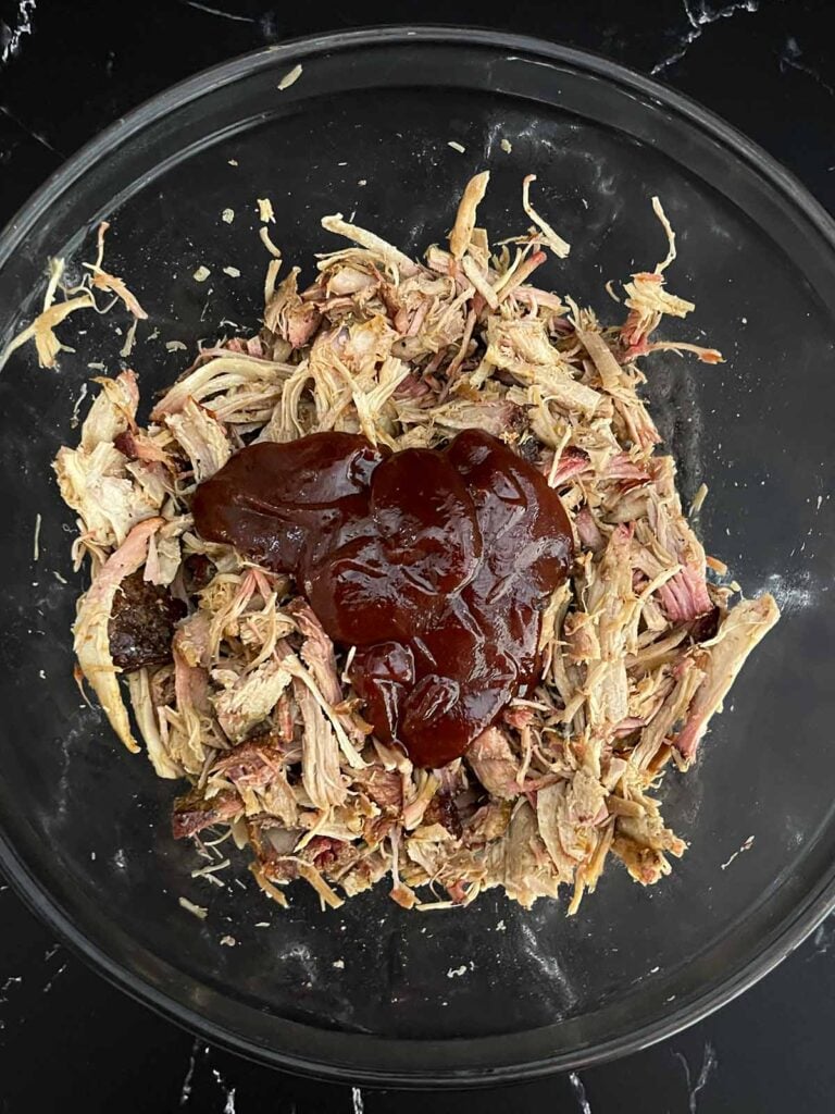Barbecue sauce poured over smoked pulled pork in a mixing bowl.