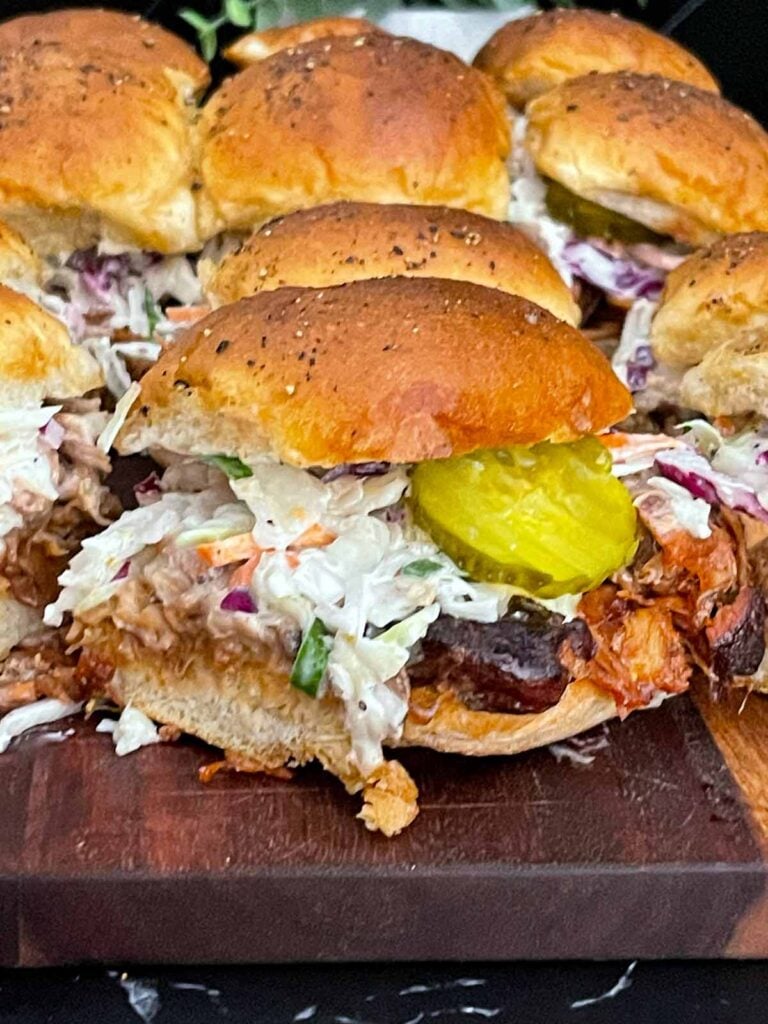 Finished pulled pork sliders assembled with coleslaw and dill pickles on savory rolls.