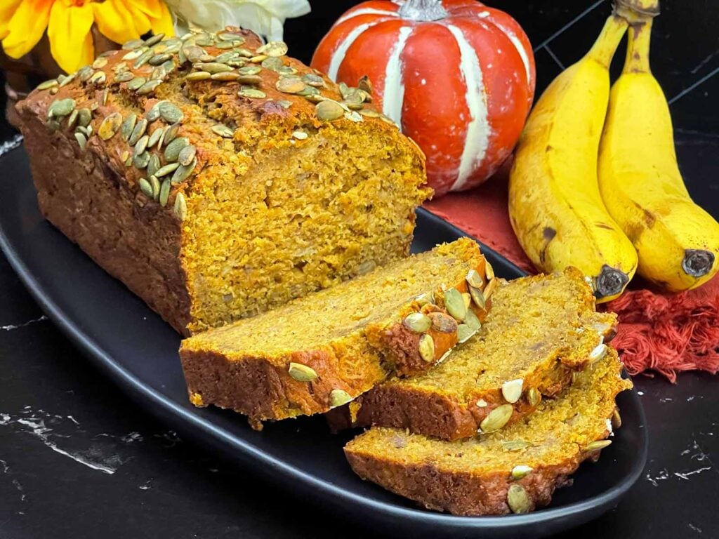 Sliced pumpkin banana bread topped with pumpkin seeds on a black platter.