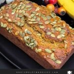 Freshly baked pumpkin banana bread topped with pumpkin seeds on a black platter.