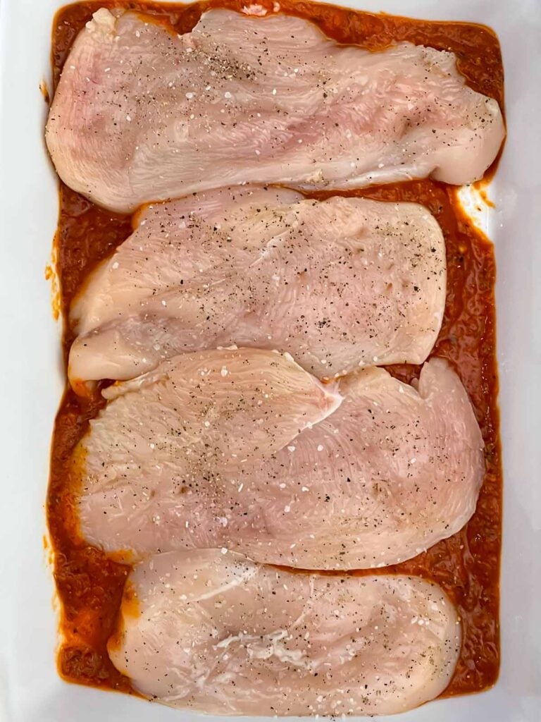 Seasoned chicken breasts arranged over marinara sauce in a baking dish.