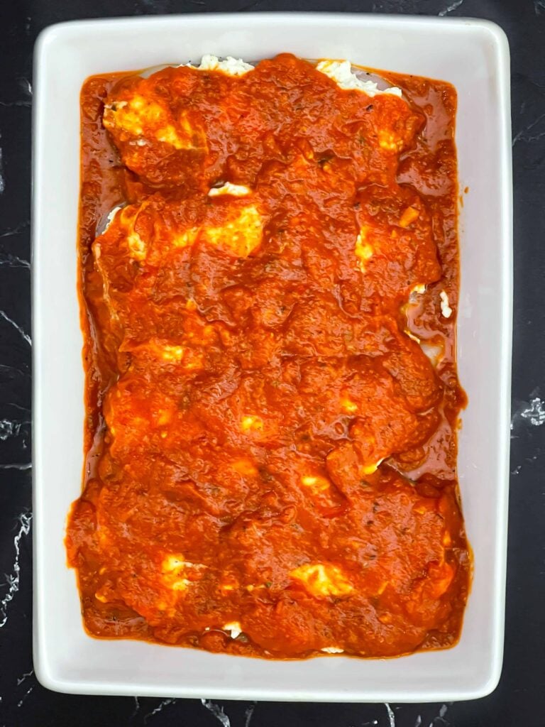 Ricotta topped chicken breasts covered with marinara sauce in a baking dish.