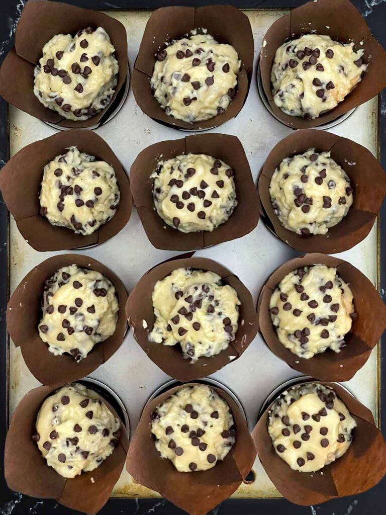 Bakery style chocolate chip muffin batter scooped into paper-lined muffin cups topped with extra chocolate chips.