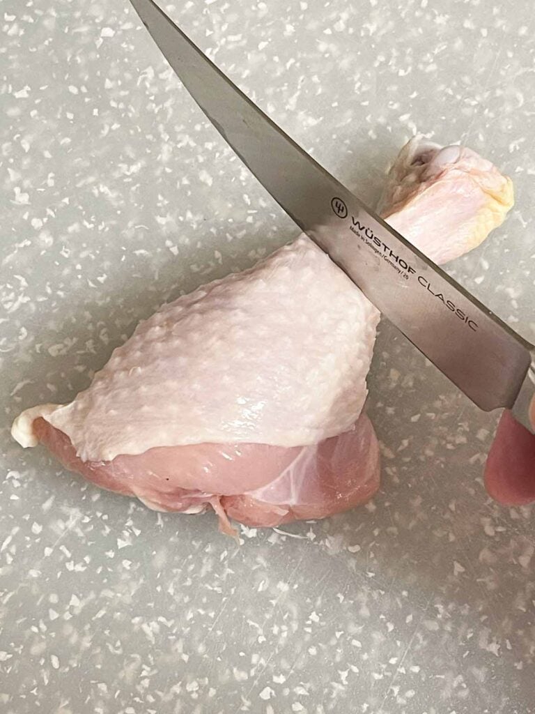 A knife cutting around the skinny end of a chicken drumstick to begin making a chicken lollipop.