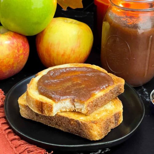 Slow cooker apple butter in a mason jar surrounded by fresh apples and smeared over a piece of sourdough toast.