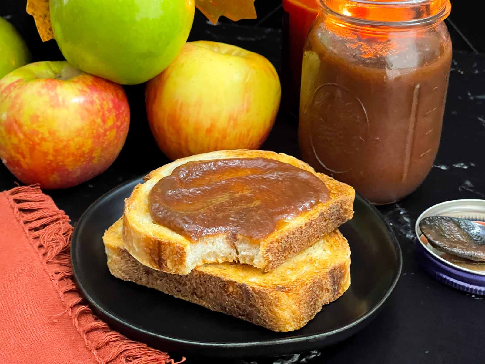 Slow cooker apple butter in a mason jar surrounded by fresh apples and smeared over a piece of sourdough toast.